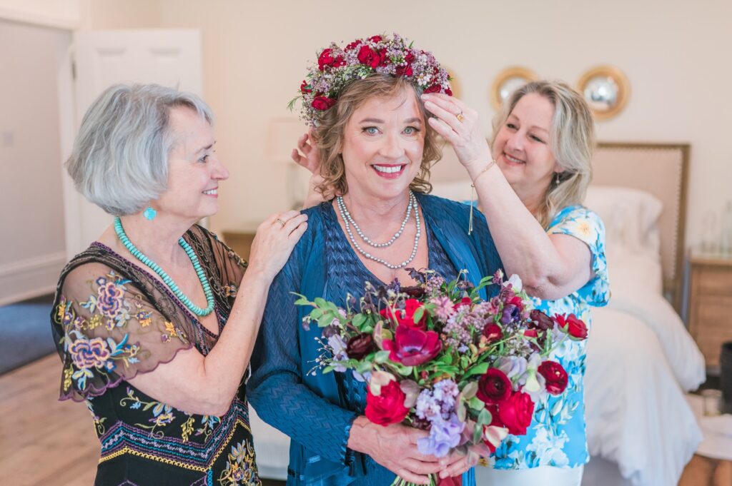 Three women standing together in a bedroom; two women adjust the floral headpiece of the smiling woman holding a bouquet in the center.
