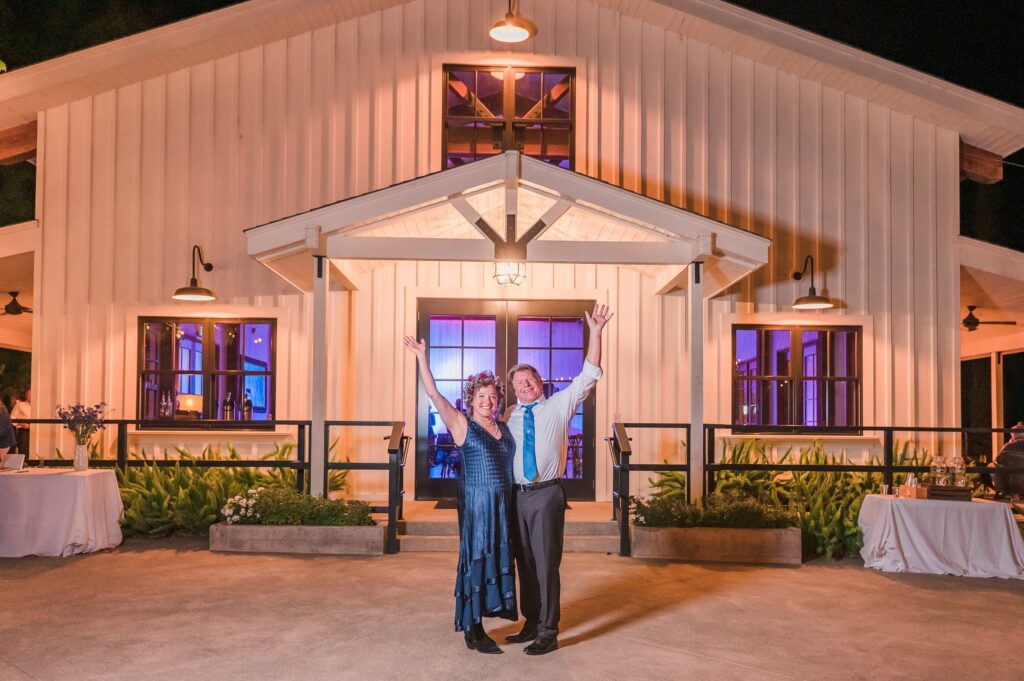 A couple stands with raised arms in front of a brightly lit barn-style venue at night, surrounded by tables and plants.