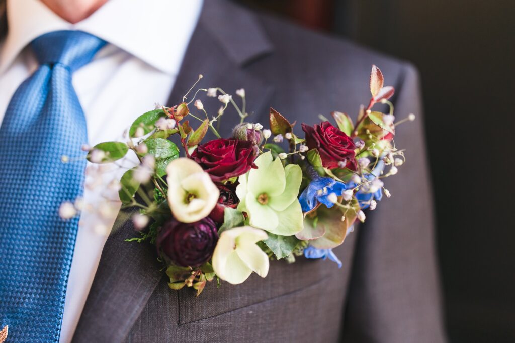 A close-up of a man's suit with a vibrant floral boutonniere featuring red, green, and blue flowers tucked in the lapel pocket.