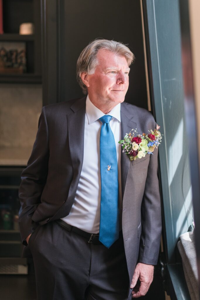 A man in a suit with a blue tie and a floral boutonniere stands by a window, looking outside.