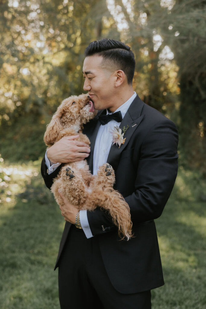 A man in a suit holds a small dog in a grassy area. The dog licks the man's face affectionately.