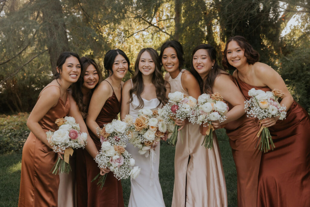 A bride and six bridesmaids in coordinated dresses pose outdoors, holding floral bouquets.
