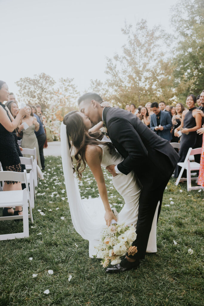 A bride and groom kiss while the groom dips the bride after their outdoor wedding ceremony, surrounded by guests who are clapping.