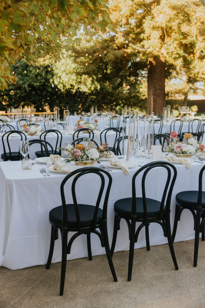 Outdoor dining setup with white tablecloths, black chairs, floral centerpieces, and candles under trees with sunlight filtering through leaves.