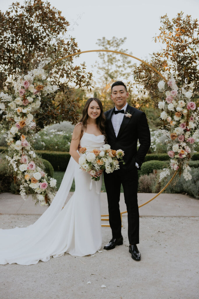 Bride and groom stand together outdoors, both smiling. The bride holds a bouquet, and a large floral wreath decorates the background.