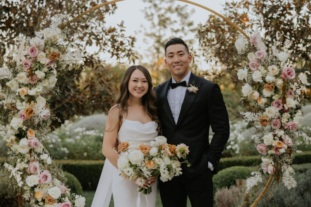 A couple stands together under a floral arch, smiling. The woman holds a bouquet of flowers. The setting appears to be an outdoor garden.