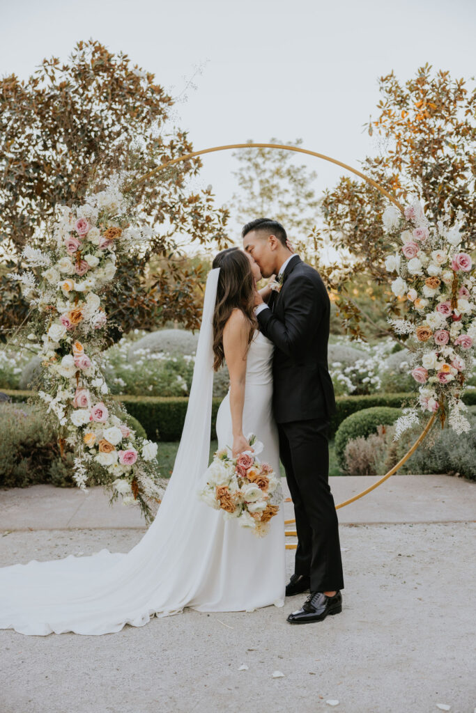 A bride and groom share a kiss in front of a circular floral arch. The bride holds a bouquet, and both are dressed in wedding attire.