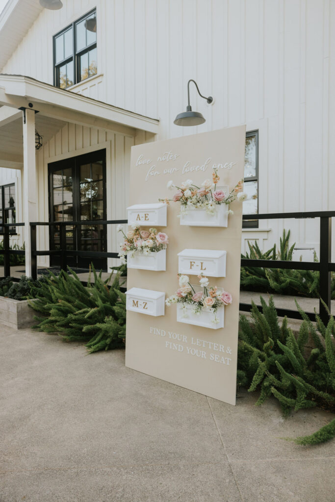 Wedding seating arrangement board with floral decorations, displaying guest letters A-E, F-L, and M-Z in white boxes. The board is in front of a white building with large windows.