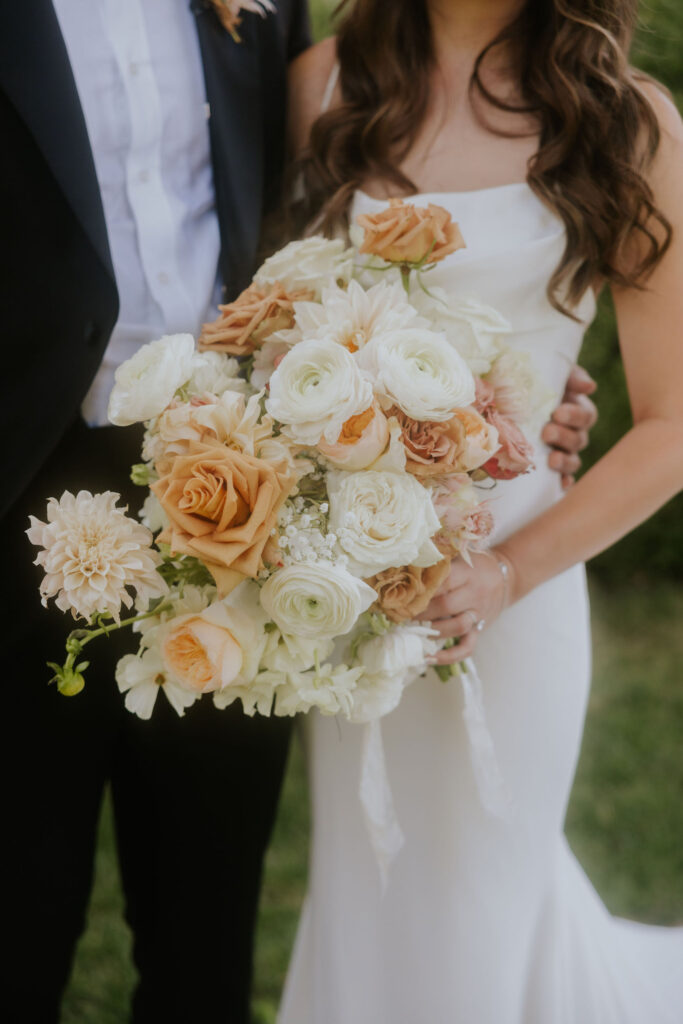 A couple stands together, with the woman holding a large bouquet of white, peach, and orange flowers.