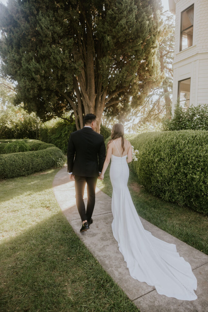 A couple in formal attire walks hand in hand on a garden path, next to green hedges and trees.