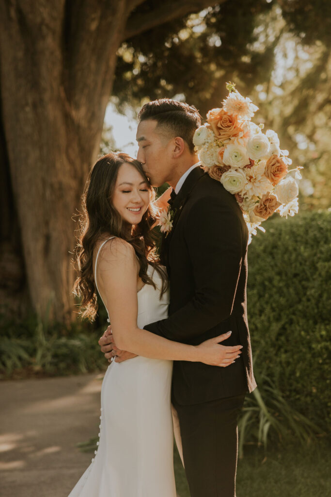 A couple embraces outdoors. The man kisses the woman's forehead while holding a bouquet of roses. They both appear happy.