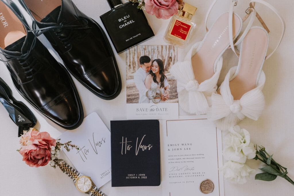 Flat lay of wedding items: black shoes, perfume, heels, a photo card, vow booklets, and an invitation surrounded by flowers and a watch.