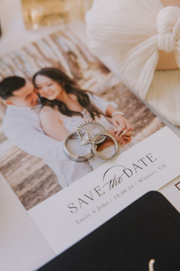 Close-up of a save-the-date card with a smiling couple and two rings on top, reading "Save the Date: Emily & John, 10.08.22, Winters, CA.