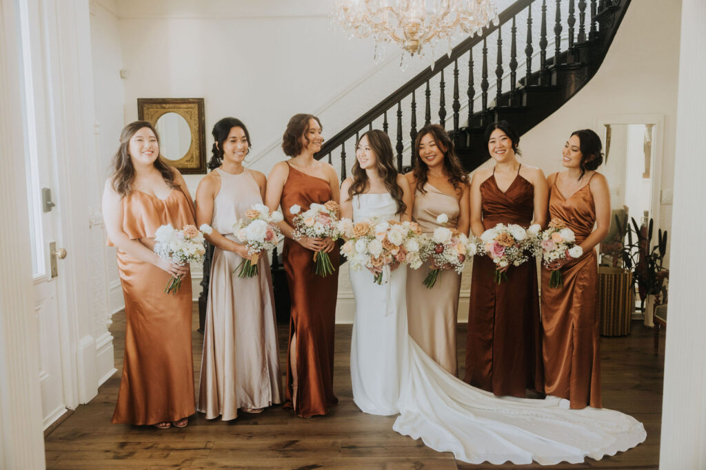 A bride stands with six bridesmaids in elegant dresses, holding bouquets, in a room with a chandelier and staircase.