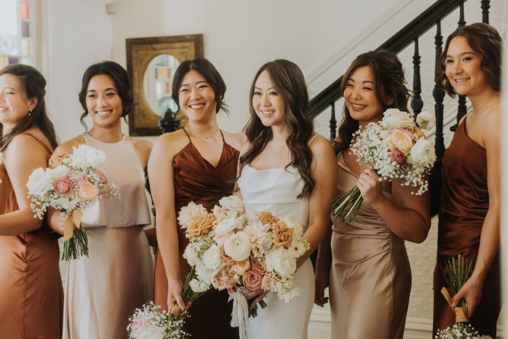 A bride in a white dress stands with five bridesmaids in various shades of brown and blush dresses, holding bouquets of flowers, in a well-lit room near a staircase.