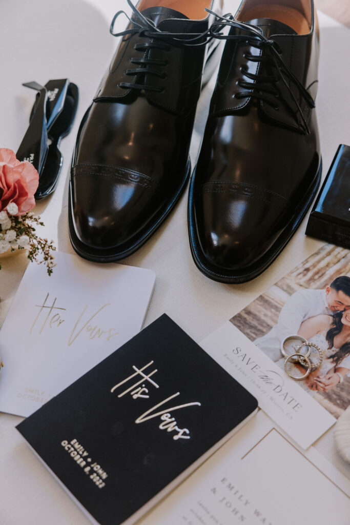 Black formal shoes, a "Her Vows" booklet, wedding invitations, a pair of rings, and a boutonniere arranged on a white surface.