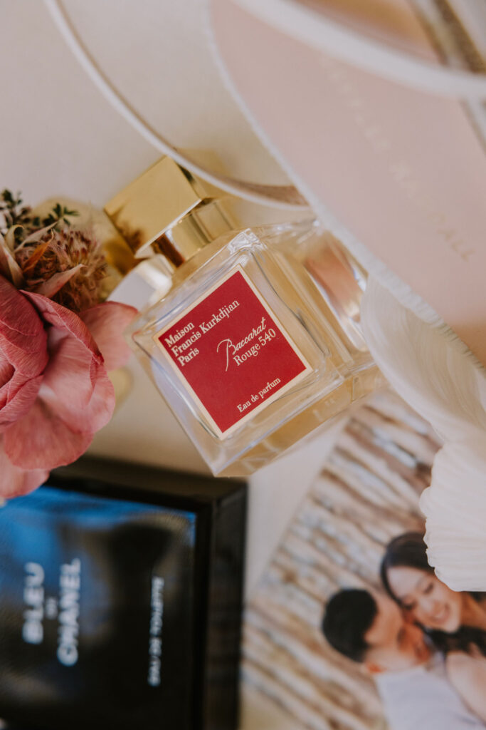 A bottle of Maison Francis Kurkdjian Baccarat Rouge 540 perfume is placed on a flat surface with pink flowers, a black Chanel box, and a photo in the background.