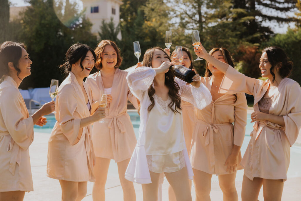 A group of women in matching robes celebrate by a pool, one drinking from a champagne bottle while others hold glasses, smiling and laughing in the sunlight.