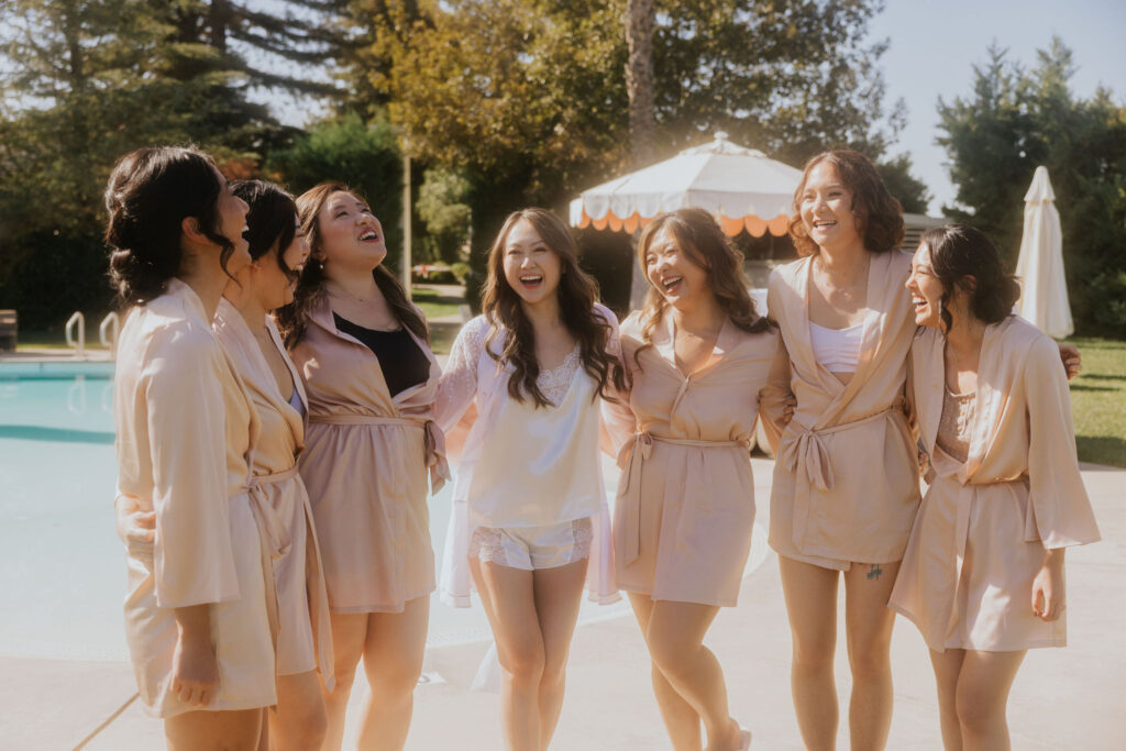 Seven women in robes stand together, smiling and laughing near a pool on a sunny day. Trees and umbrellas are in the background.