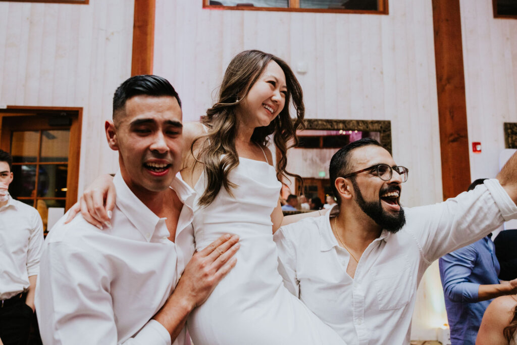 A woman in a white dress is lifted by two men in a festive indoor setting. Everyone appears to be having fun.
