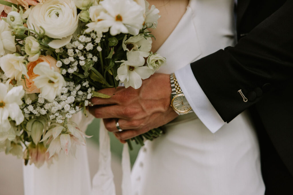 Close-up of a couple holding a bouquet of white and peach flowers. The person on the right wears a black suit and a watch.