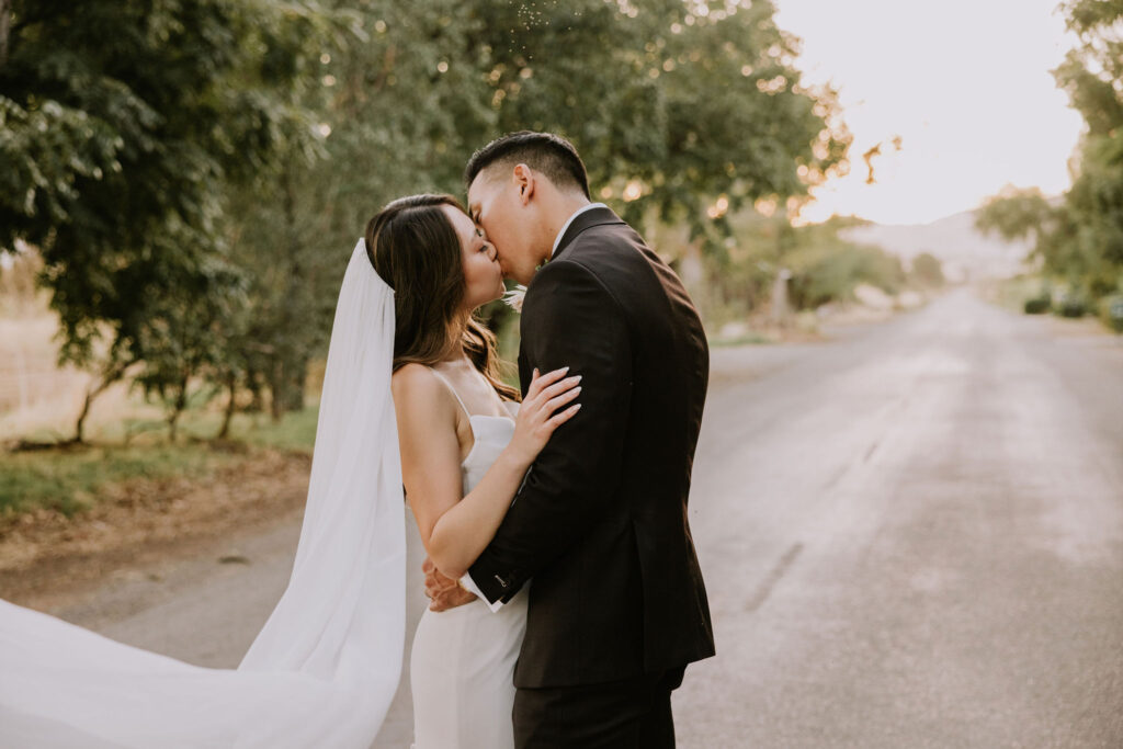 A couple in wedding attire shares a kiss on a tree-lined road at sunset.