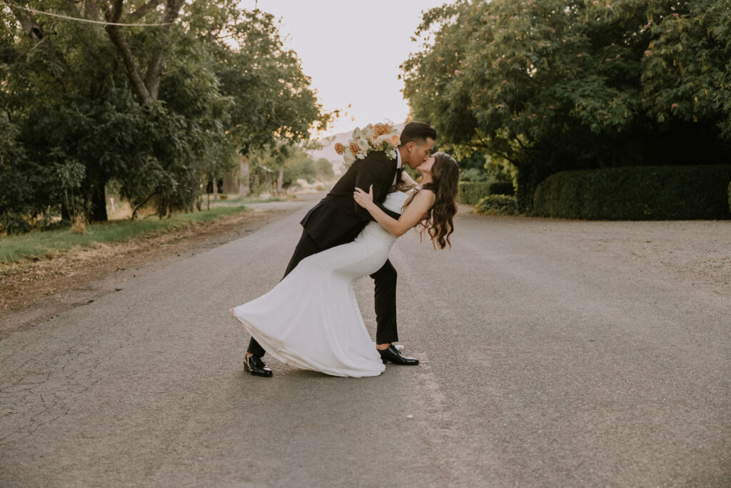 A couple in formal attire embraces in a dip on a tree-lined road, with the woman holding a bouquet of flowers.