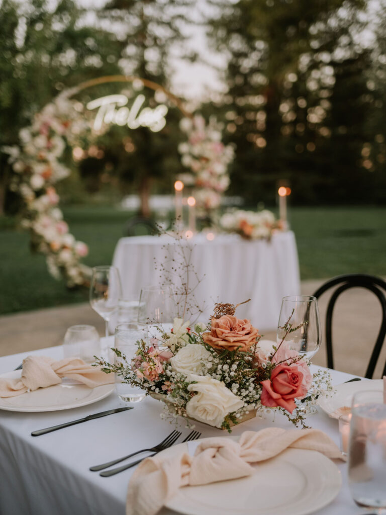 Elegant outdoor wedding table setting with floral centerpiece, candles, and a decorative arch in the background.