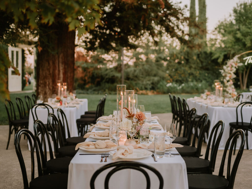Elegant outdoor wedding setup with round tables covered in white tablecloths, black chairs, floral centerpieces, and tall candle arrangements in a lush garden setting.