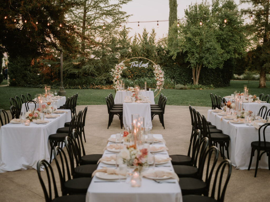 Outdoor wedding reception setup with long tables covered in white tablecloths, black chairs, floral centerpieces, and a decorative arch with the text "The Lees" in the background.