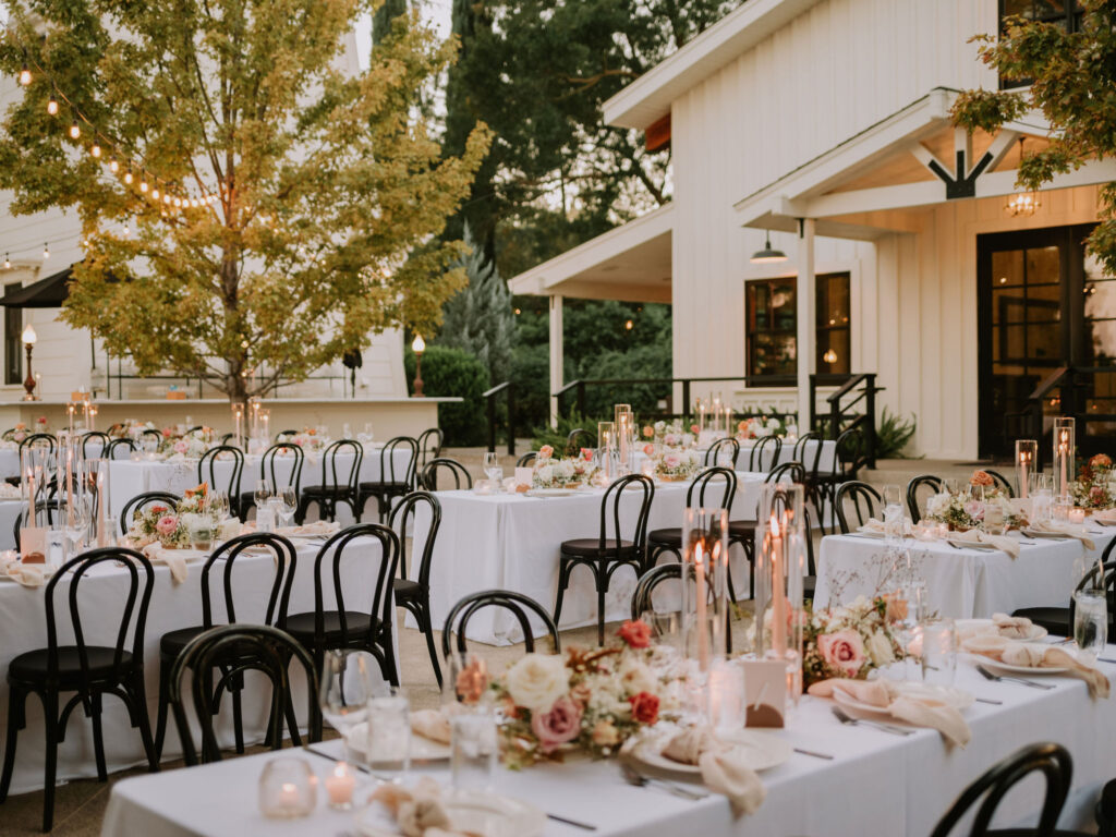 Outdoor wedding reception with elegantly set tables, white tablecloths, floral centerpieces, and black chairs. String lights hang above, creating a warm ambiance near a white building and trees.