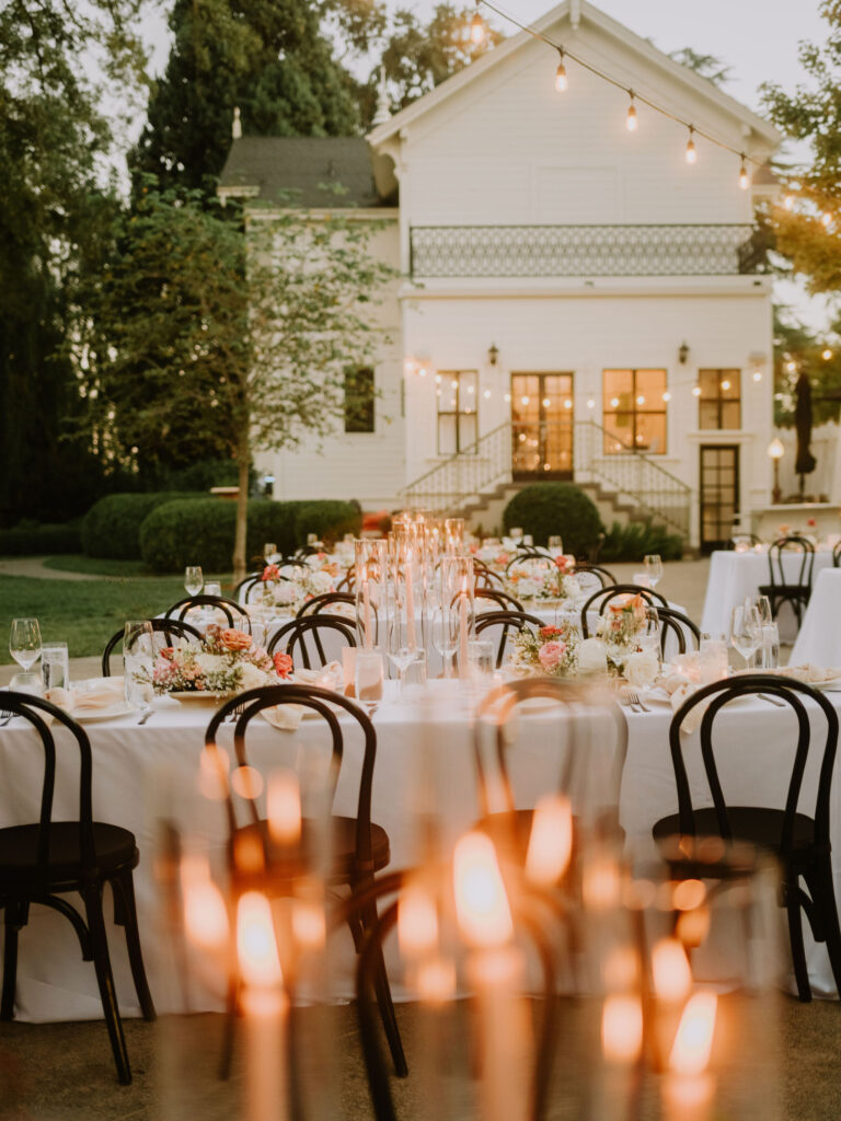 Outdoor wedding reception setup with long tables, white tablecloths, floral centerpieces, candles, and black chairs. A white house and string lights are visible in the background.