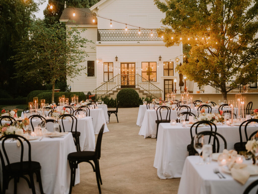 Outdoor wedding reception with tables covered in white linens, floral centerpieces, and lit candles. String lights hang above, and a large house is in the background.