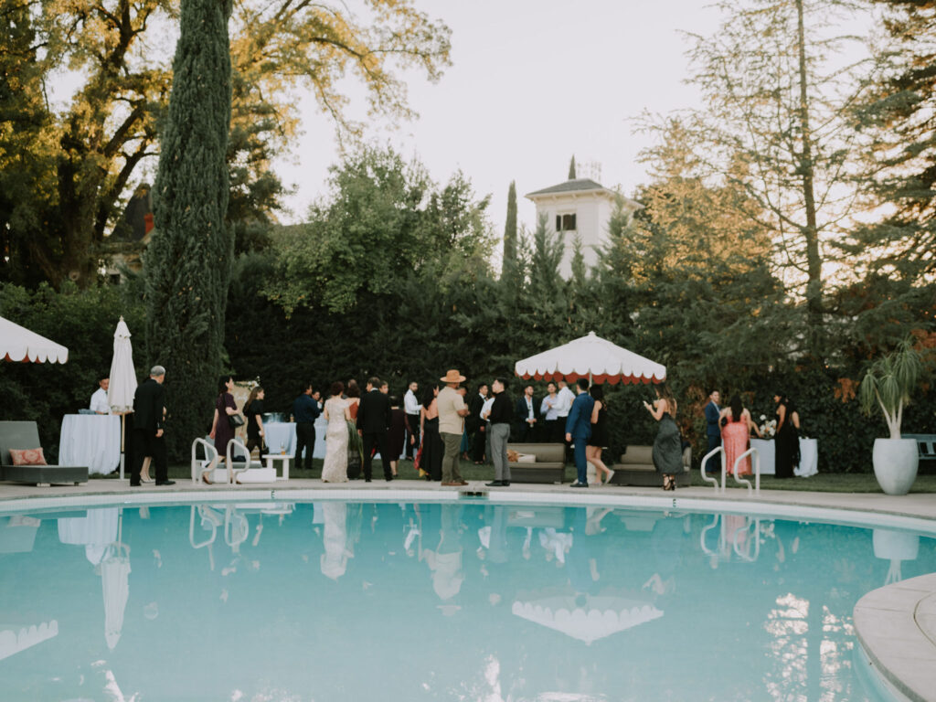 People gathered around a pool with trees and a building in the background on a clear day.