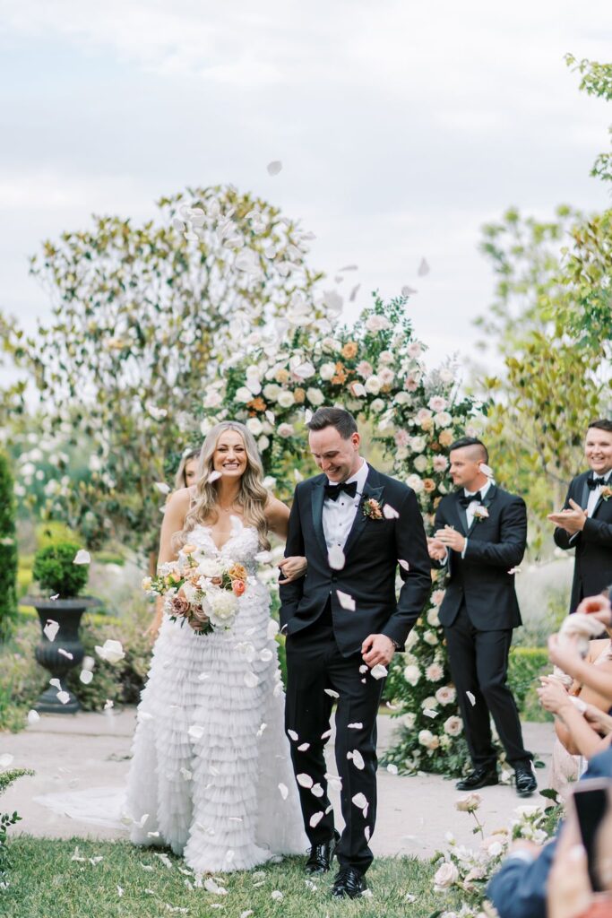 A bride and groom walk down the aisle outdoors, surrounded by guests throwing flower petals. An arch decorated with flowers is in the background.