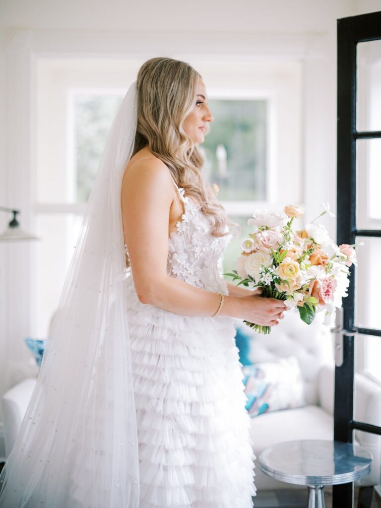 Bride in a white gown holding a bouquet stands by a window, with long veil and tiered dress facing left.