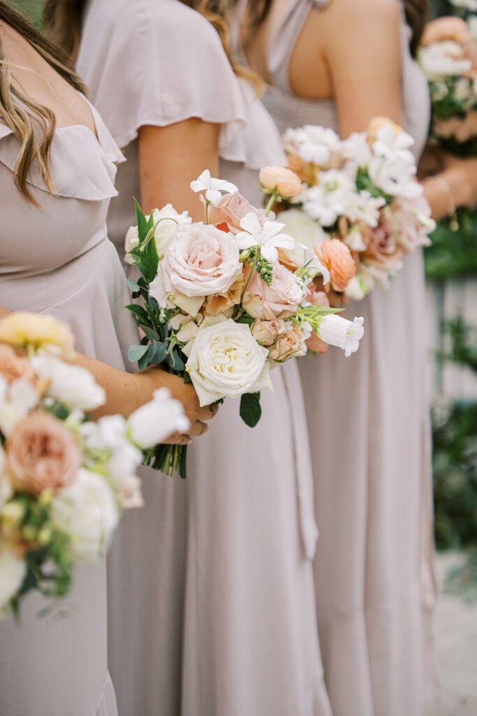 Bridesmaids in light dresses holding bouquets of roses and other flowers.