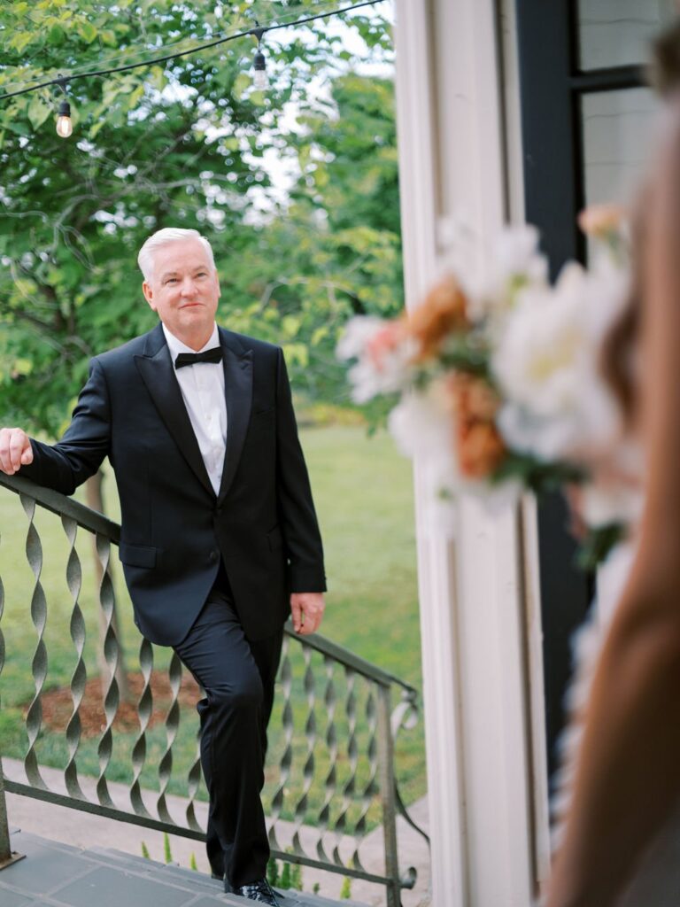 Man in a black tuxedo stands on stairs, holding a railing, with a blurred bouquet in the foreground. Outdoor greenery is visible in the background.