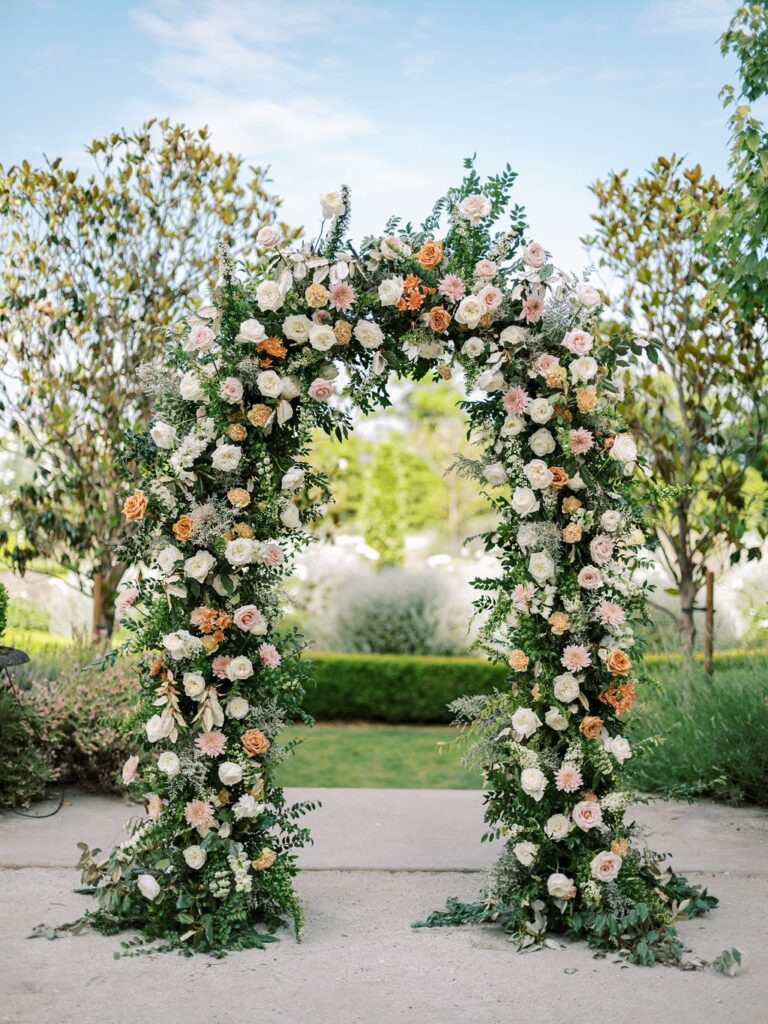 An archway decorated with lush green foliage and pastel roses, set outdoors with trees and a garden visible in the background.