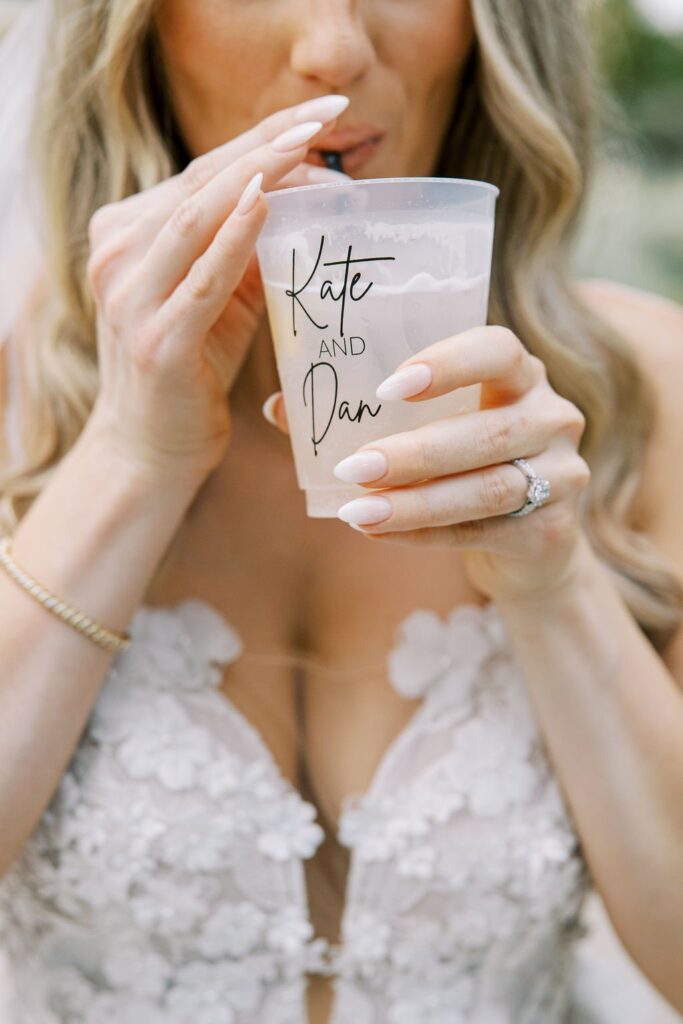 Woman in a white floral dress drinking from a clear cup with "Kate and Dan" written on it.