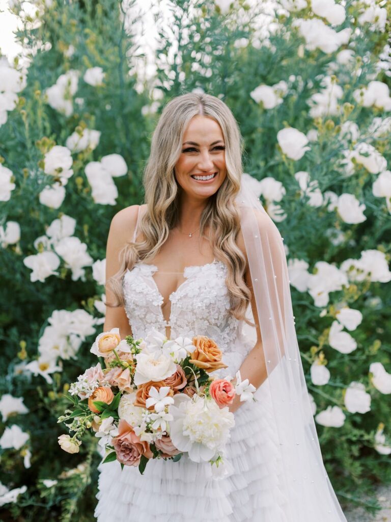 A bride with long wavy hair smiles, holding a bouquet of white, orange, and pink flowers. She stands in front of a backdrop of white flowering plants, wearing a detailed white wedding dress.
