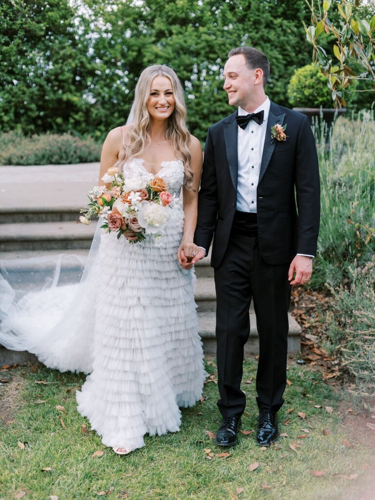 A bride in a white dress and a groom in a black tuxedo hold hands and smile in a garden setting. She holds a bouquet of flowers.