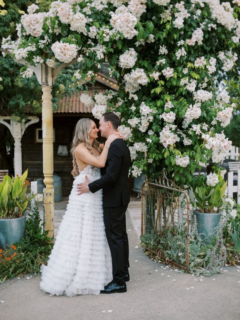 A bride and groom embrace under an arch covered with white flowers in an outdoor setting.