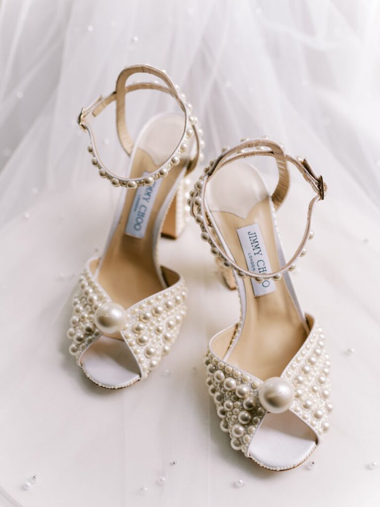 Beige high-heeled sandals adorned with pearl embellishments and ankle straps, set on a white fabric background.