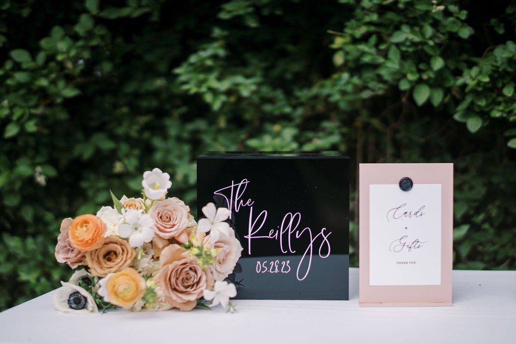 A black box with "The Kellys 05.08.23" next to a card and floral arrangement, set against green foliage.