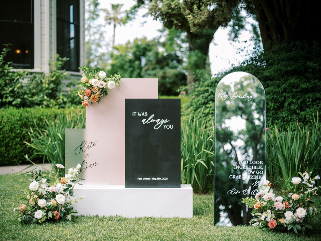 Floral signs outdoors read "IT WAS always YOU" and "Hey Cutie, you look incredible. Now go grab a drink." Surrounded by lush greenery and floral arrangements.
