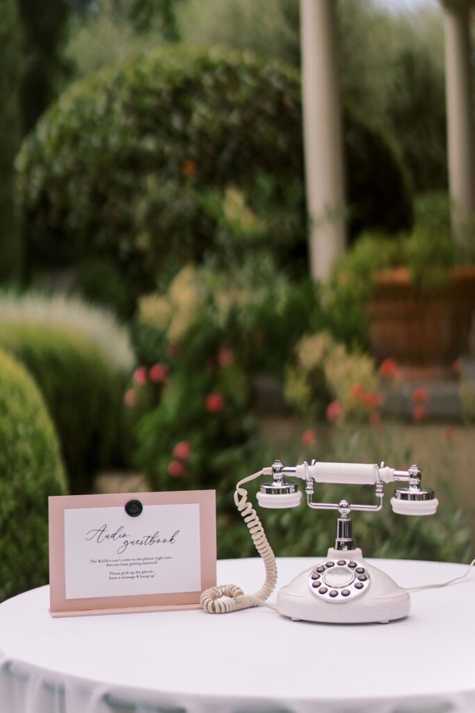 A vintage rotary telephone sits on a table outdoors next to a guestbook sign, with plants and a columned structure in the background.