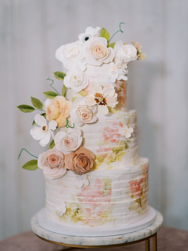 A three-tiered wedding cake with pastel floral decorations and leaves, placed on a round marble stand.