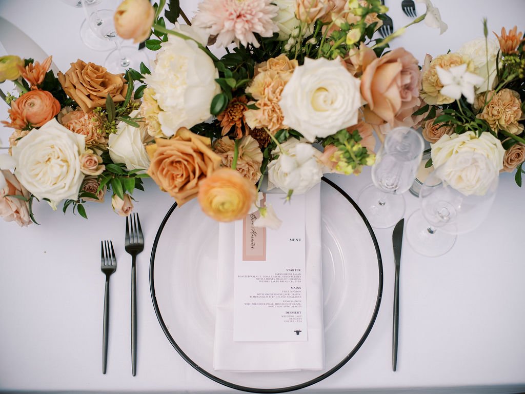 Elegant table setting with floral arrangements in peach and white tones, featuring a menu placed on a white plate with black cutlery on either side.