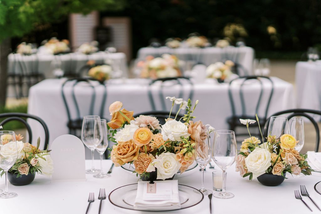 Elegant outdoor dining setup with white tablecloths, floral centerpieces featuring roses, and neatly arranged cutlery and glassware. Black chairs surround the tables.